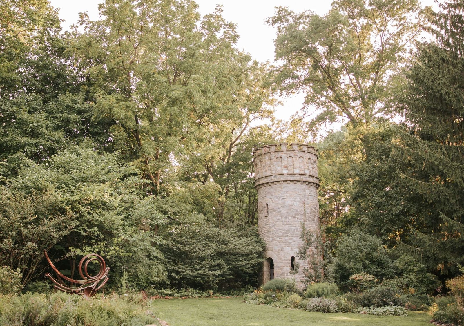 A round stone tower standing among tall summer trees, with a lawn and a curling metal sculpture in the foreground.
